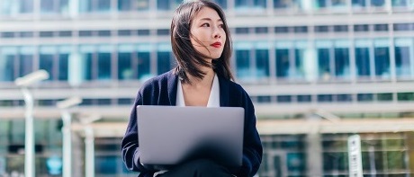 Eine junge Geschäftsfrau sitzt vor einem Bürokomplex auf einer kleinen Mauer mit Laptop auf dem Schoß. Sie schaut nach rechts als blicke sie in die Zukunft als Freelancerin.