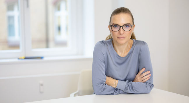 Tatjana Kiel sitzt mit verschränkten Armen am Tisch und schaut in die Kamera